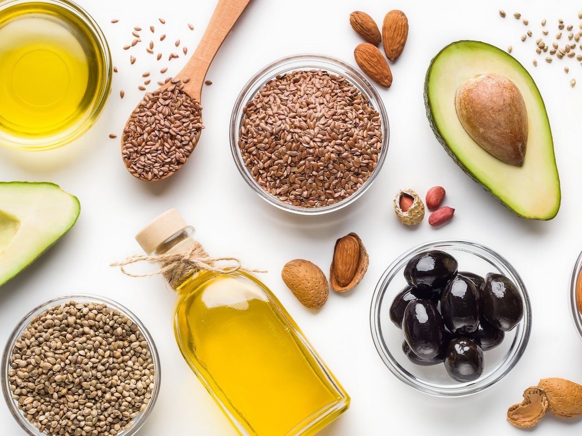Avokado, keten tohumu, zeytinyağı, kenevir tohumu, badem ve siyah zeytin gibi çeşitli sağlıklı yağ kaynakları, beyaz bir zemin üzerinde sergileniyor.