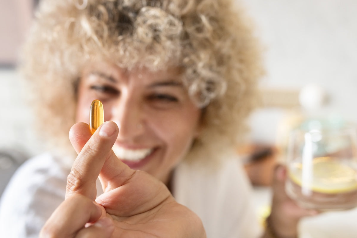 Parmaklarının arasında altın renkli bir omega-3 takviye kapsülü tutan gülümseyen kadın; arka planda bir bardak su bulunuyor. Günlük sağlık ve zindeliği teşvik eden bir sahne.