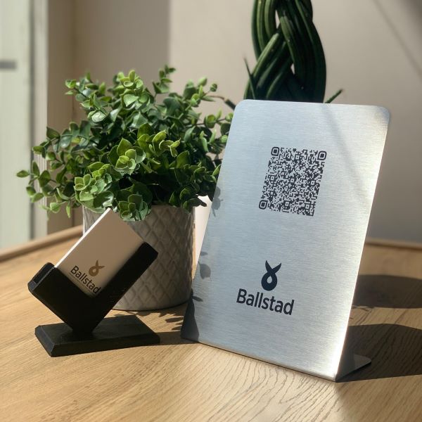 Bir masada Ballstad logolu metal QR kod tabelası ve kartlık, arka planda dekoratif yeşil bitkiler.