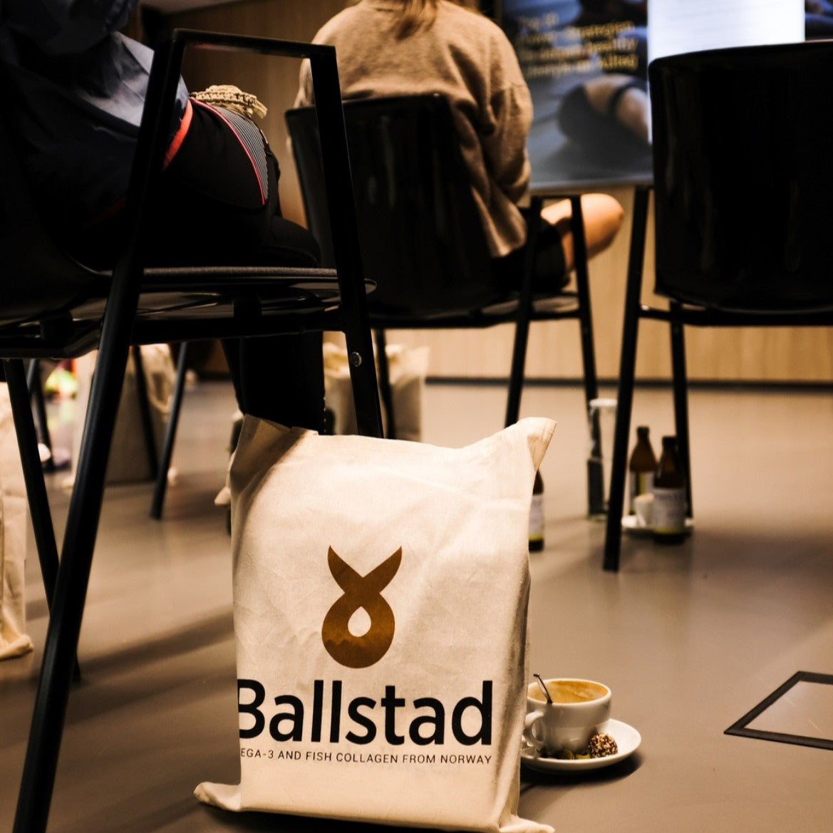 Ballstad logolu bez çanta, bir seminer veya etkinlik sırasında sandalyelerin altında yer alıyor; yanında kahve fincanı ve atıştırmalık bulunuyor.