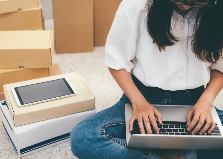 Beyaz gömlek giymiş bir kadın, yerde oturmuş dizüstü bilgisayarda çalışıyor; etrafında karton kutular ve birinin üzerinde duran tablet dikkat çekiyor. Görsel, e-ticaret veya evden çalışma temasını yansıtıyor.