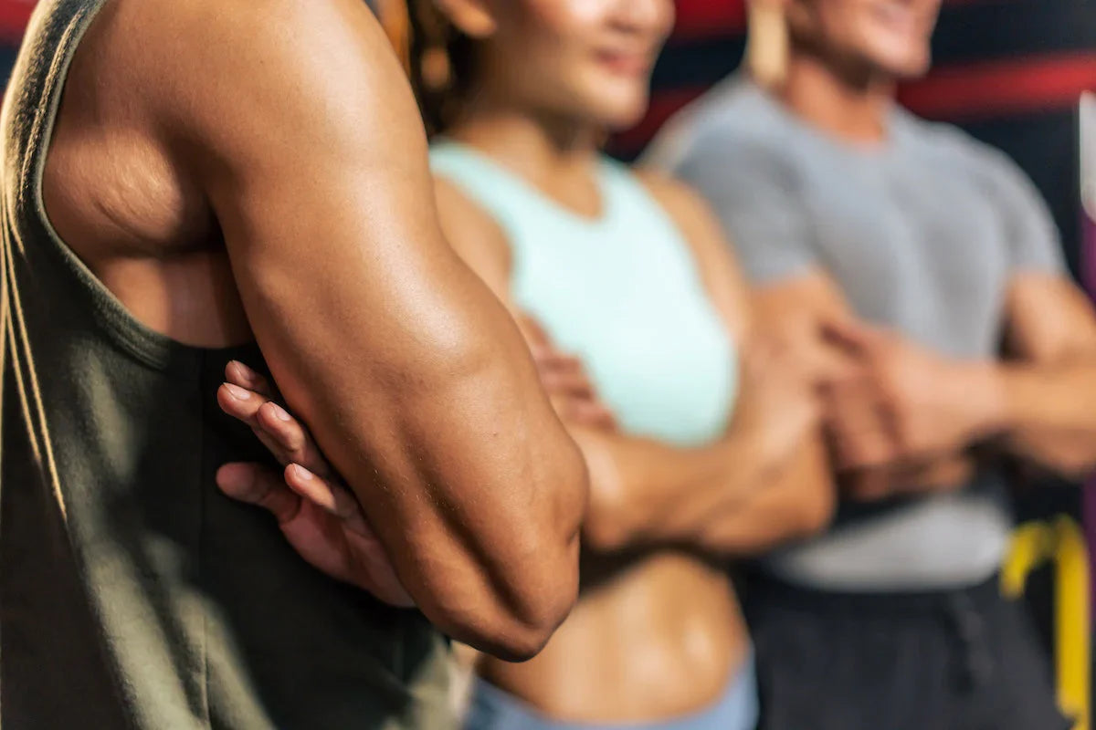 Spor salonunda kollarını göğsünde birleştirmiş, spor kıyafetleri giymiş genç kadın ve erkeklerden oluşan grup, güçlü kolları ve fit görünümleriyle yakın plan.