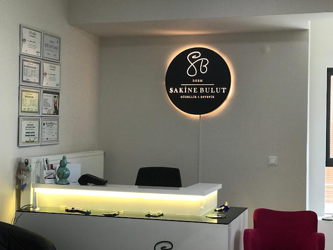 Sakine Bulut Güzellik ve Estetik merkezinin resepsiyon alanı. Duvarda aydınlatmalı yuvarlak bir logo ve sertifikaların yer aldığı bir pano bulunuyor. Beyaz banko üzerinde dekoratif objeler ve bir telefon yer alıyor, sağda pembe bir koltuk görülüyor.