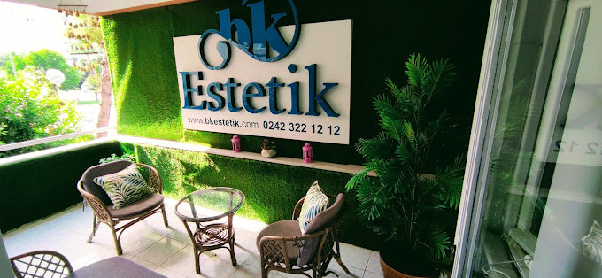 bk Estetik adlı güzellik merkezinin dış balkon alanı; yapay çim kaplı duvar, büyük tabela, cam masa, minderli sandalyeler ve dekoratif bitkilerle şık bir şekilde dekore edilmiş. Tabela üzerinde iletişim bilgileri ve web sitesi yer alıyor.
