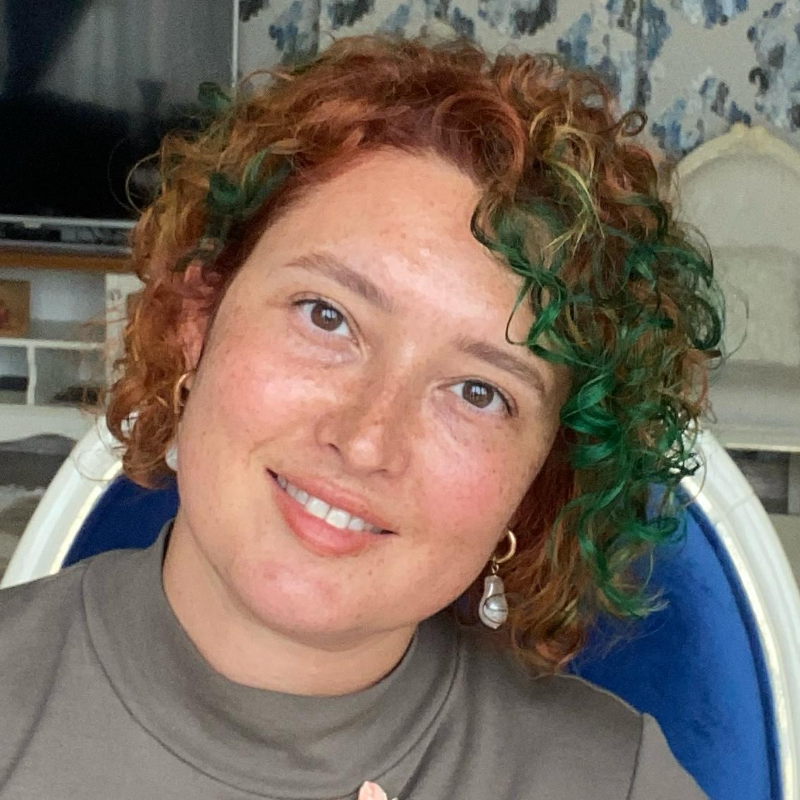 Kıvırcık ve renkli (yeşil ve kızıl tonlarında) saçlara sahip, gülümseyen bir kadın portresi. Kadın inci küpe takıyor ve nötr tonlarda bir bluz giymiş. Arka planda iç mekân detayları ve mavi bir sandalye görünüyor.