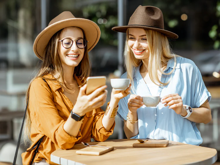 Güneşli bir günde açık hava kafede oturan iki genç kadın, kahvelerini yudumlarken cep telefonuna bakarak gülümsüyor. Şapka ve modern kıyafetlerle stil sahibi görünüyorlar, samimi bir sohbet ortamı dikkat çekiyor.