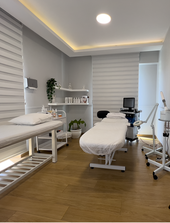 Modern ve minimalist şekilde döşenmiş bir cilt bakım odası; iki adet beyaz örtülü yatak, duvar raflarında kozmetik ürünler, bir bakım cihazı, büyüteçli lamba ve ahşap zemin üzerinde düzenli bir yerleşim ile hijyenik bir ortam sunulmuş.