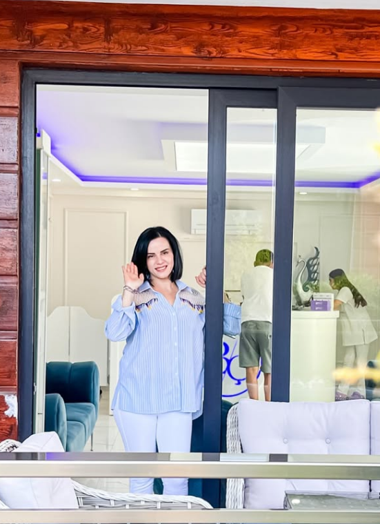 Cam kapının önünde duran ve gülümseyerek el sallayan, mavi çizgili gömlek ve beyaz pantolon giymiş bir kadın. Arka planda BC logosunun bulunduğu modern bir iç mekanda çalışan iki kişi ve mor LED aydınlatmalı tavan detayları yer alıyor.