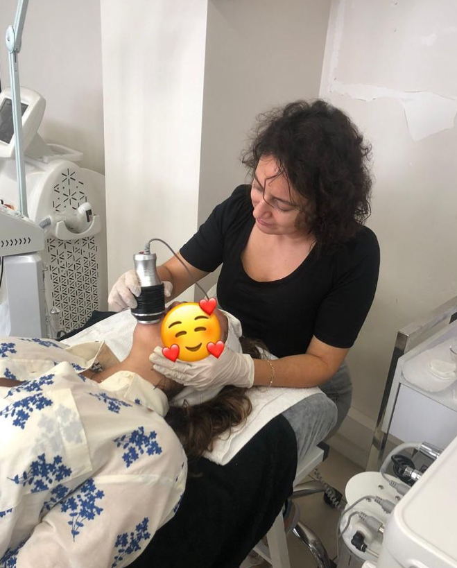 Siyah tişörtlü bir uzman, eldiven takarak bir kadına profesyonel cilt bakım cihazıyla uygulama yapıyor. Yatmakta olan müşterinin yüzü emojiyle kapatılmış, ortamda medikal estetik ekipmanları bulunuyor.