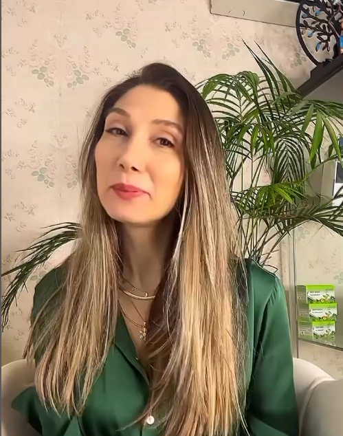 Yeşil gömlek giymiş, uzun saçlı bir kadın iç mekânda, arka planda yeşil bitki ve birkaç kutu ürünle birlikte kameraya gülümsüyor.