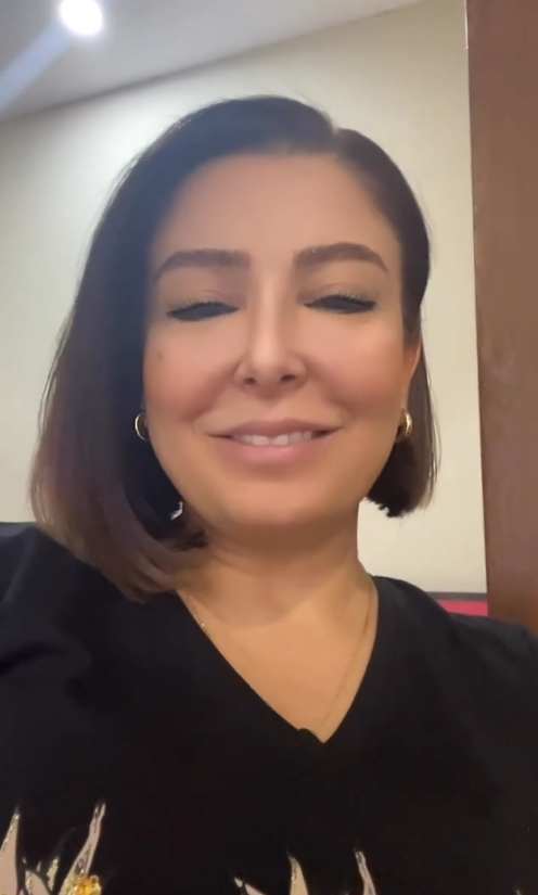 Kısa kahverengi saçlı, altın halka küpeler takan ve siyah desenli bir bluz giymiş bir kadın, kapalı mekânda gülümseyerek kameraya bakıyor.