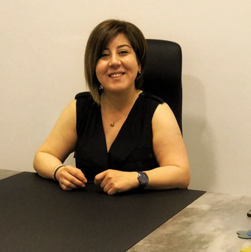 Ofis ortamında, siyah kolsuz bluz giymiş ve masada oturan, gülümseyen bir kadın. Arka planda sade bir duvar ve siyah bir ofis sandalyesi görülüyor.