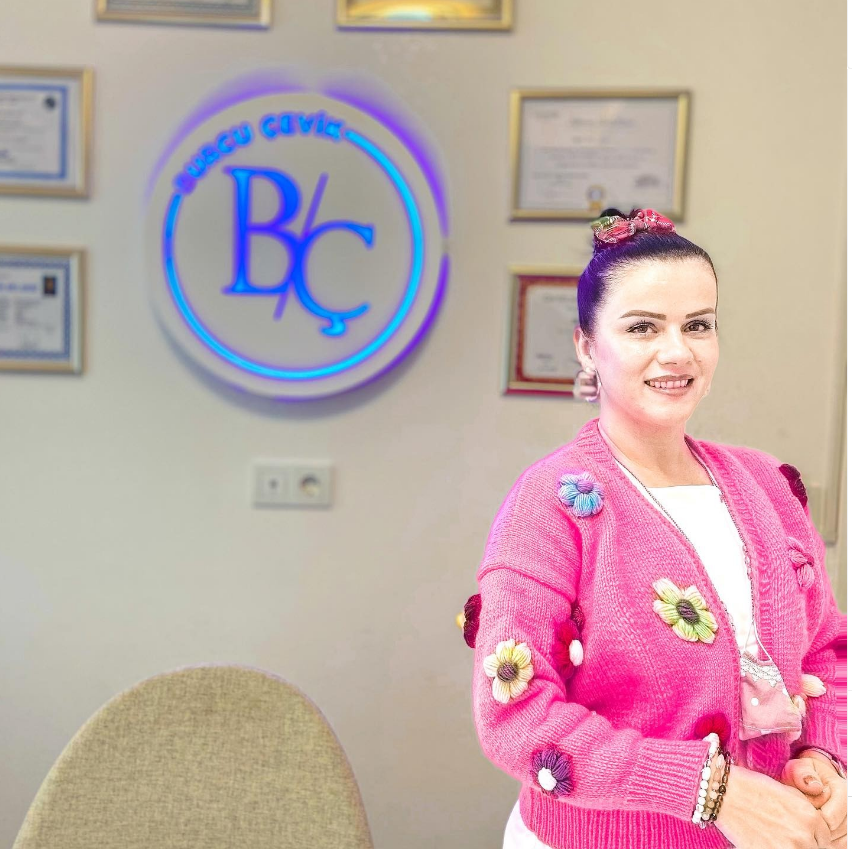 Burcu Çevik logosunun önünde pembe çiçekli hırka giymiş, saçları yukarıdan toplu ve güler yüzlü bir kadın. Arka planda çerçeveli sertifikalar ve mavi ışıklı BC logosu yer alıyor.