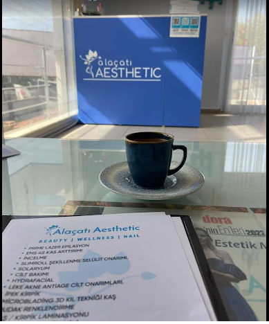 Alaçatı Aesthetic merkezinde, cam bir masa üzerinde duran mavi fincanda bir kahve; arka planda mavi renkli resepsiyon bankosu üzerinde 'Alaçatı Aesthetic' logosu yer alıyor. Masada bir hizmet broşürü ve bir moda/estetik dergisi bulunmakta.