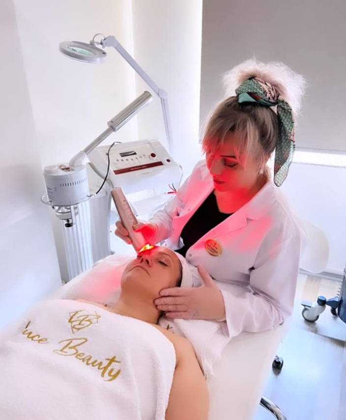 Beyaz önlük giymiş bir uzman, estetik yatağında uzanan kadının alnına kırmızı ışık yayan bir cihazla cilt bakımı uyguluyor. Kadının üstünde 'Luce Beauty' yazılı havlu bulunmakta, başında bone var. Arka planda profesyonel bakım ekipmanları yer almakta.