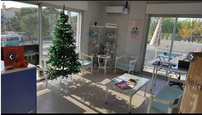 Alaçatı Aesthetic merkezinin bekleme alanında, pencere önüne yerleştirilmiş süslenmiş bir yılbaşı ağacı, cam vitrinde sergilenen cilt bakım ürünleri ve beyaz-mavi tonlarında klasik tarzda oturma grubu yer almakta. Masalarda dergiler, broşürler ve çalışma evrakları bulunurken, dışarıda mavi masa-sandalye takımları dikkat çekiyor.