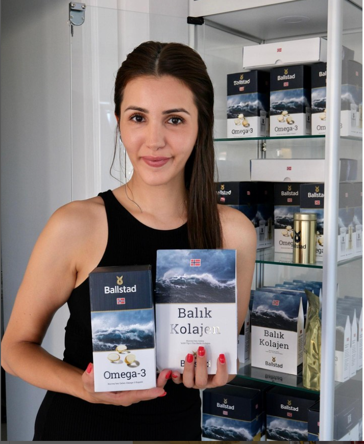 Siyah elbise giymiş genç bir kadın, elinde Ballstad marka Omega-3 ve Balık Kolajeni ürün kutularını tutarak kameraya gülümsüyor. Arka planda cam raflı dolapta aynı markaya ait ürünler sergileniyor.