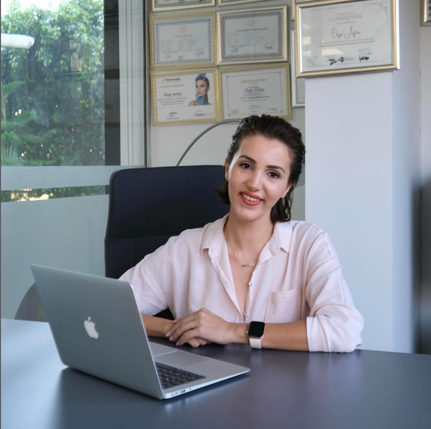 Açık pembe gömlek giymiş genç bir kadın, ofis ortamında bir masada MacBook bilgisayarının başında gülümseyerek poz veriyor. Arka duvarda çeşitli eğitim ve başarı sertifikaları asılı; camın dışından yeşillikler görülüyor.