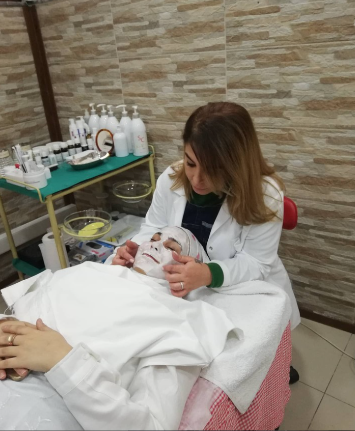 Beyaz önlük giymiş bir uzman, spa yatağında uzanan kadının yüzüne beyaz bir maske uyguladıktan sonra parmak uçlarıyla nazikçe masaj yapıyor. Kadın gözleri kapalı şekilde dinlenmekte; arka planda cilt bakım ürünlerinin bulunduğu yeşil servis arabası yer alıyor.