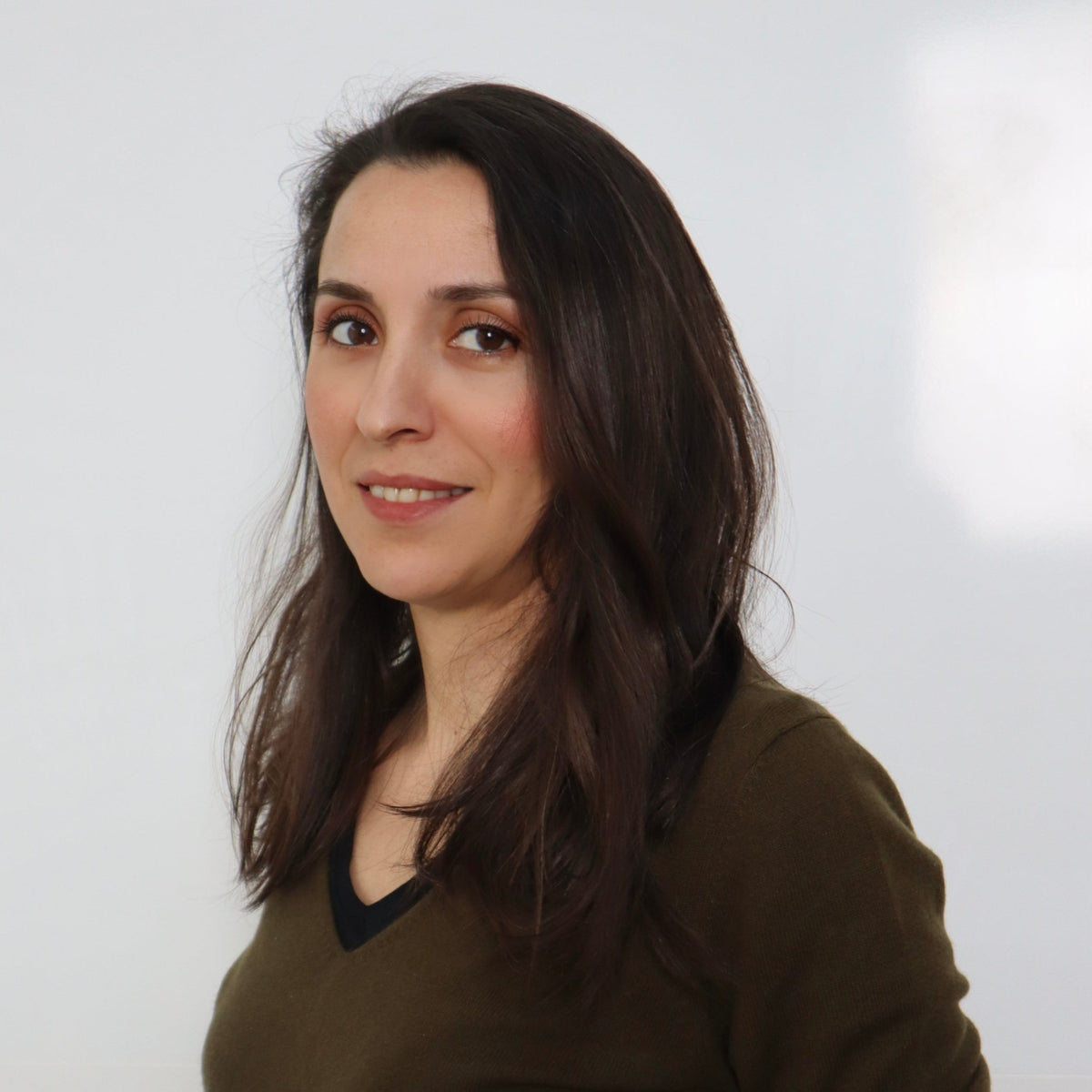 Uzun koyu kahverengi saçlı, haki renkli V yaka kazak giymiş ve hafif tebessüm eden Meryem Kulli beyaz arka plan önünde poz veriyor.