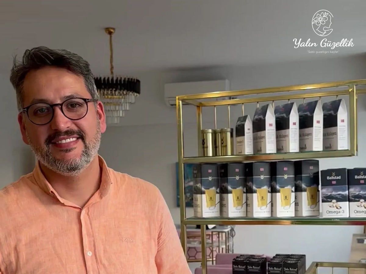 Pembe keten gömlek giymiş, gözlüklü ve gülümseyen bir adam, raflarda Ballstad marka omega-3 takviyeleri ve diğer sağlık ürünlerinin sergilendiği bir mekânda poz veriyor. Üst köşede 'Yalın Güzellik' ve 'Saklı güzelliğini keşfet' yazısı yer alıyor.