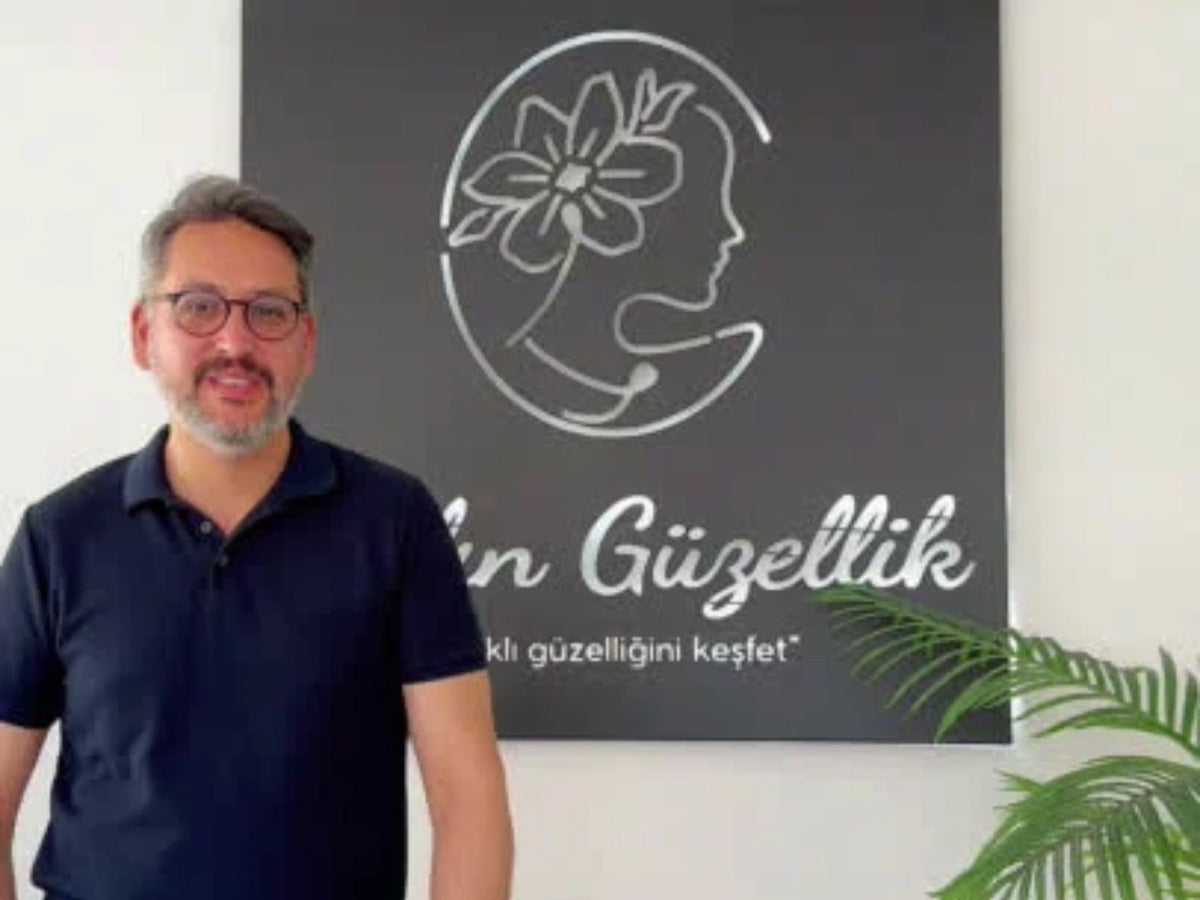 Koyu lacivert tişört giymiş gözlüklü bir adam, arkasında 'Yalın Güzellik' logosunun ve 'Saklı güzelliğini keşfet' sloganının yer aldığı siyah bir pano önünde ayakta poz veriyor. Sağ alt köşede yeşil bir bitki yaprağı görülüyor.