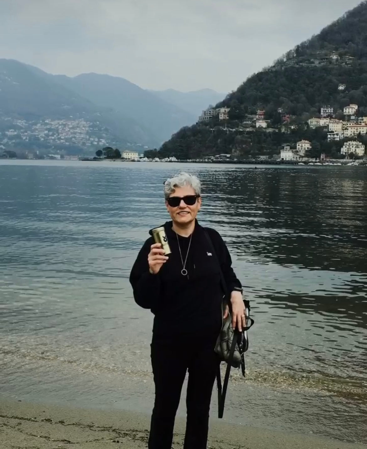 Doğal bir göl manzarası önünde elinde Ballstad altın ambalajlı kolajen kutusunu tutan, siyah kıyafetli ve beyaz saçlı kadın; doğayla iç içe sağlık ve güzellik vurgusu.