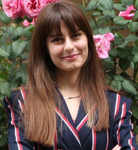 Gül çiçekleri önünde poz veren genç kadın; lacivert, bordo ve beyaz çizgili ceket giyiyor, saçları düz ve perçemli.