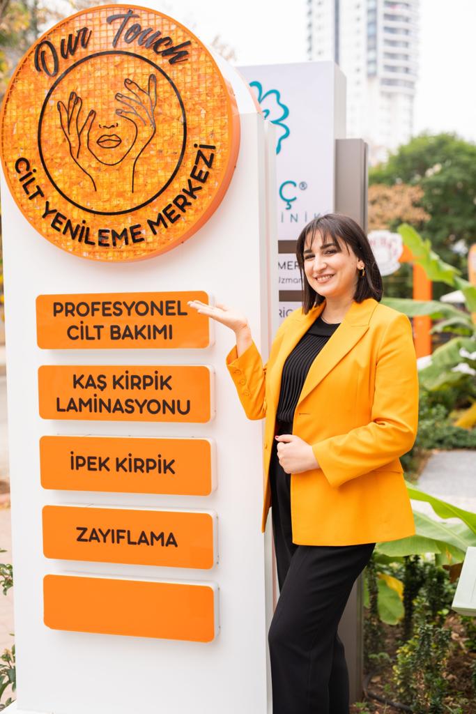 Turuncu ceketli bir kadın, Our Touch Cilt Yenileme Merkezi'nin önünde durarak 'Profesyonel Cilt Bakımı' tabelasını gösteriyor. Arka planda kaş-kirpik laminasyonu, ipek kirpik ve zayıflama hizmetleri listeleniyor.