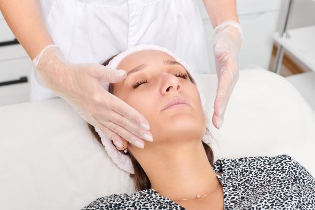 Bir kadın spa yatağında gözleri kapalı şekilde uzanmış, başında beyaz bir saç bandı var. Eldivenli bir uzman, elleriyle yüzüne nazikçe masaj yapıyor. Kadın siyah-beyaz desenli bir kıyafet giymekte.